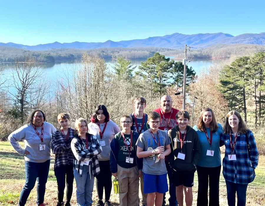 A group of youth members pose during the 2023 Confirmation Retreat. Behind them is a large body of water, mountains, and trees.