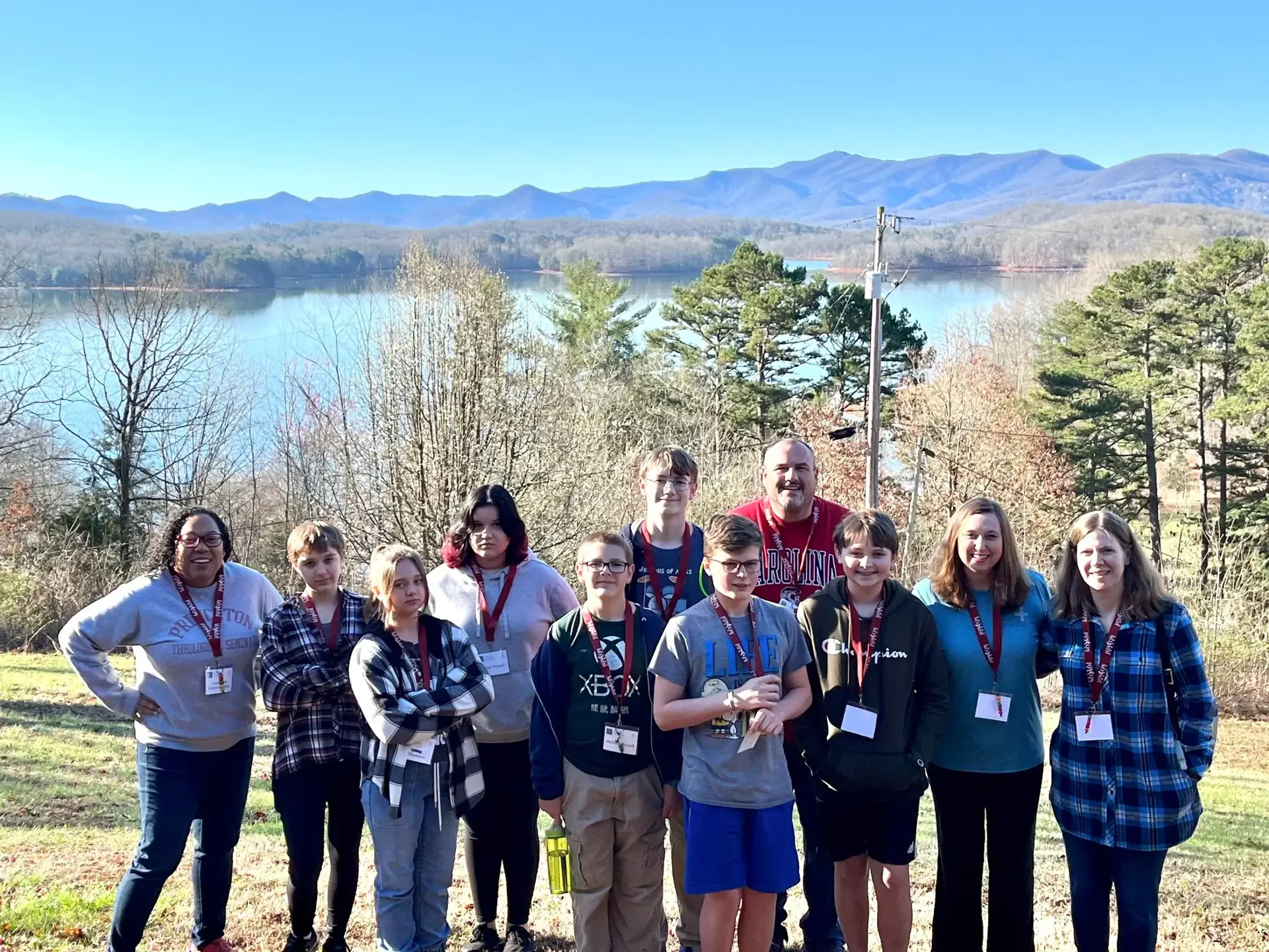 A group of youth members pose during the 2023 Confirmation Retreat. Behind them is a large body of water, mountains, and trees.