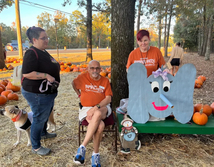 Patch volunteers sit at the Pumpkin Patch with orange shirts on. They are next to the pricing table. One volunteer is holding a cardboard elephant head.