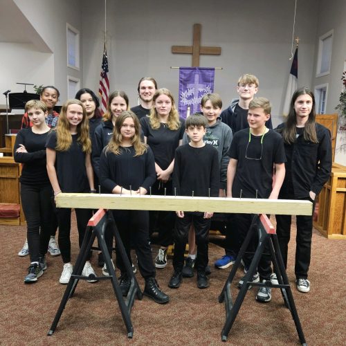 A group of youth members dressed in black stands for a picture after the Good Friday service. In front of them, there is a wooden board propped up on sawhorses with a bunch of nails in it.