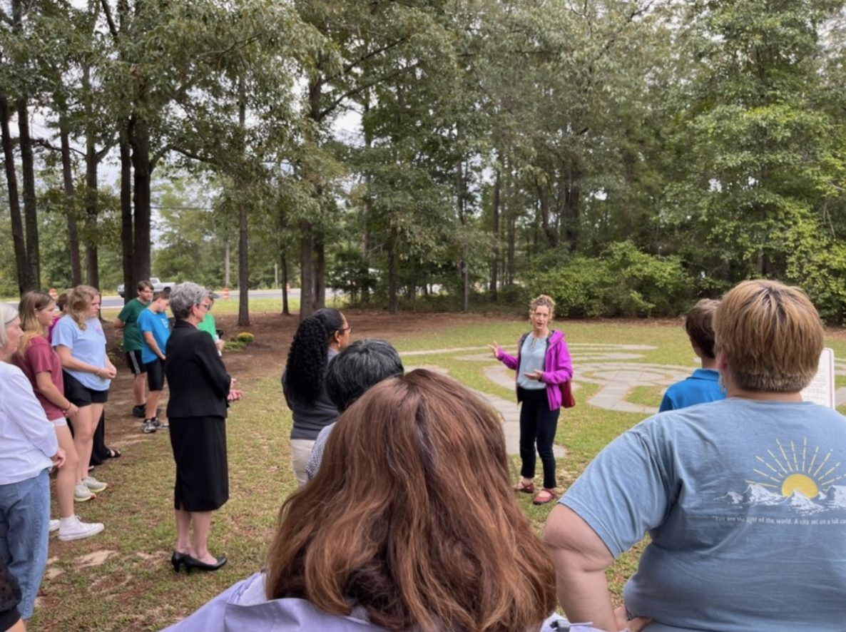 A large group of church members stands listening to a presenter during the dedication of the prayer labyrinth.